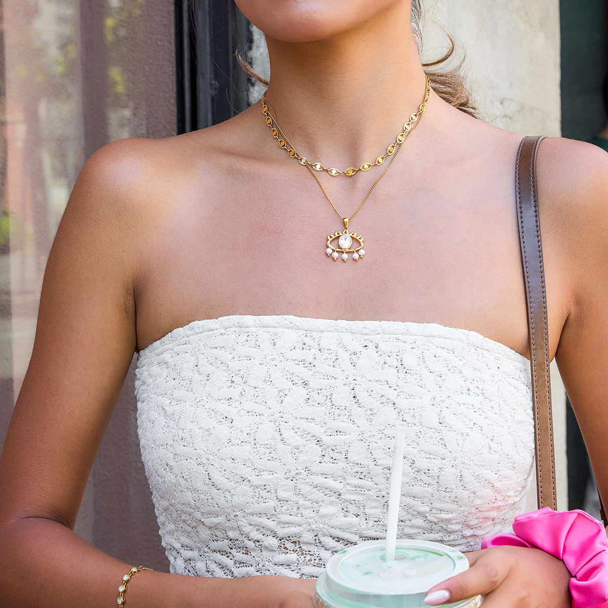 Woman wearing a gold necklace with a pendant, holding a drink, and wearing a white strapless top.