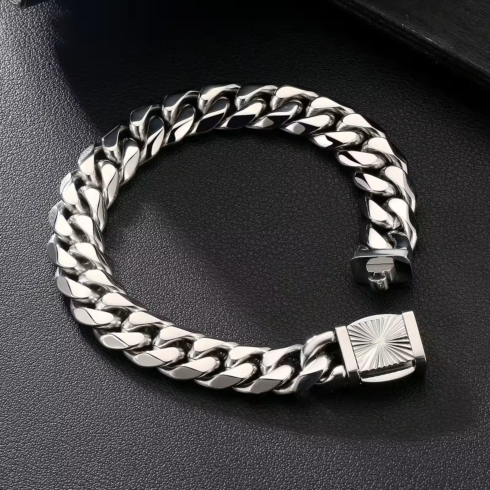 A silver-toned Cuban link chain bracelet with a square sunburst clasp, displayed on a textured black surface.