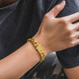 A close-up of a person's wrist wearing a chunky gold Cuban link chain bracelet with a polished rectangular clasp, paired with a dark gray t-shirt.