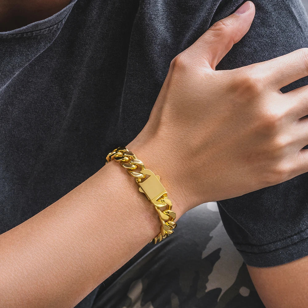 A close-up of a person's wrist wearing a chunky gold Cuban link chain bracelet with a polished rectangular clasp, paired with a dark gray t-shirt.