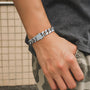 A close-up of a person's wrist wearing a chunky silver-toned metal chain bracelet with a rectangular clasp, paired with a dark gray t-shirt and camouflage pants.