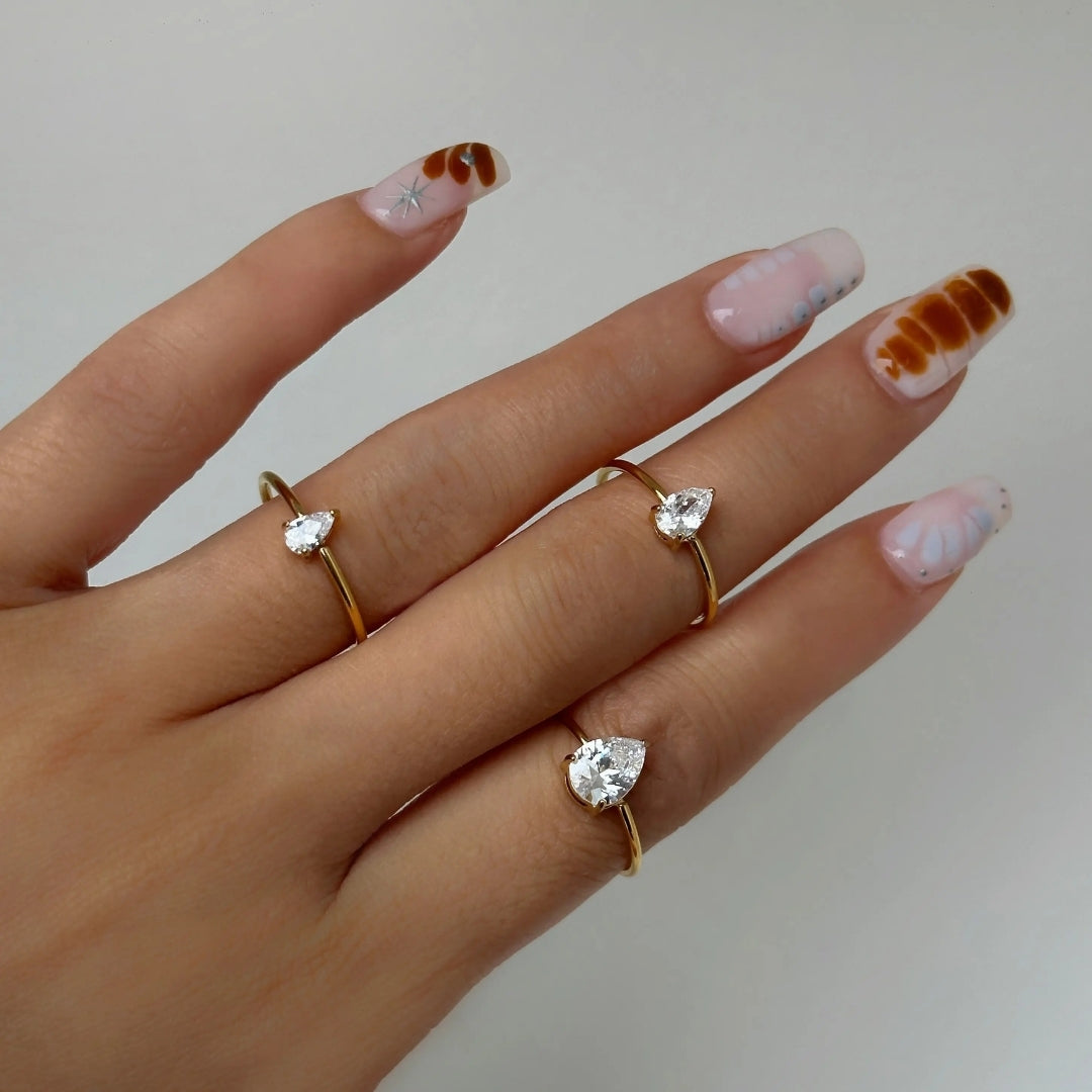Hand wearing two gold rings with heart-shaped gemstones on a neutral background