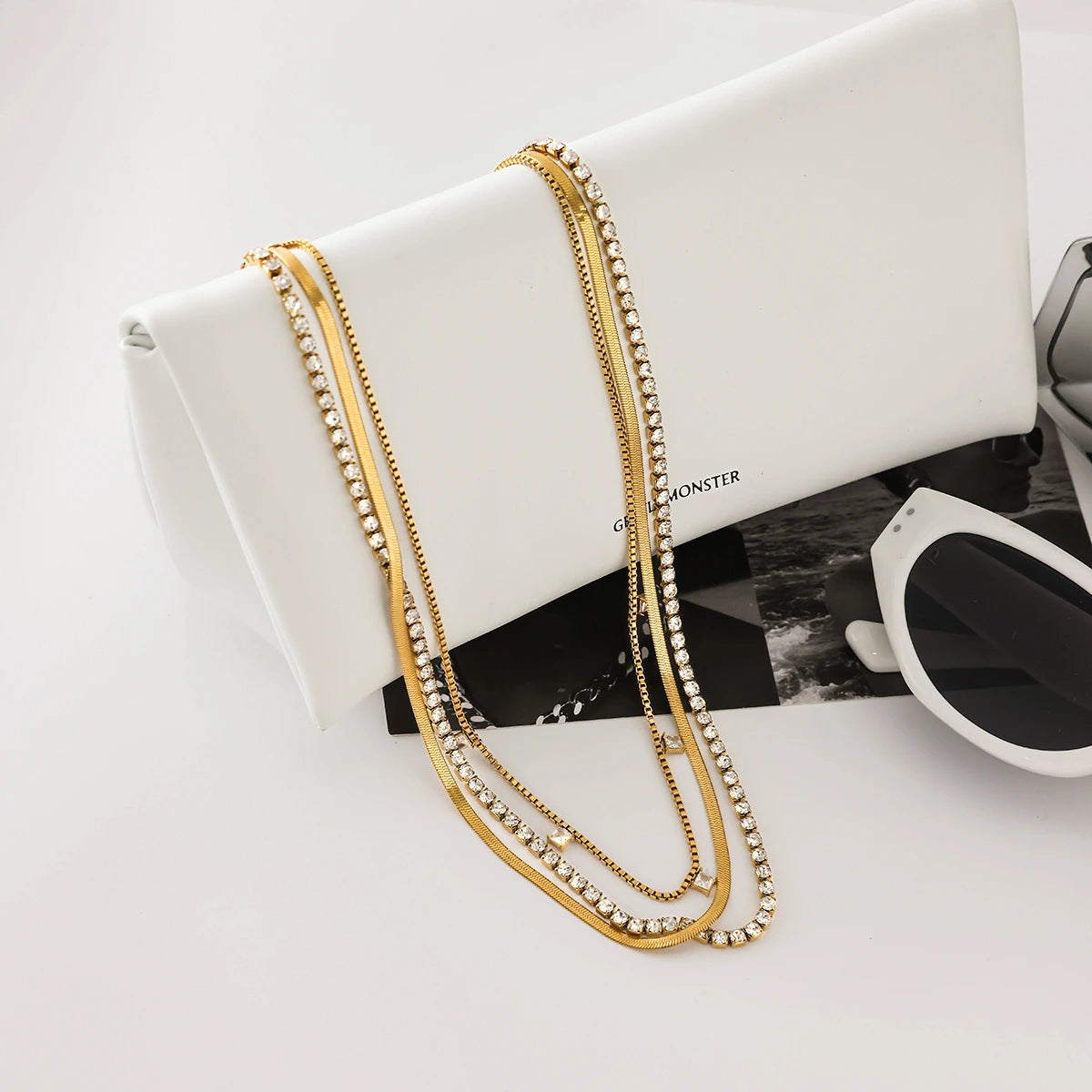 Gold chain necklace draped over a white clutch with sunglasses on a light gray background