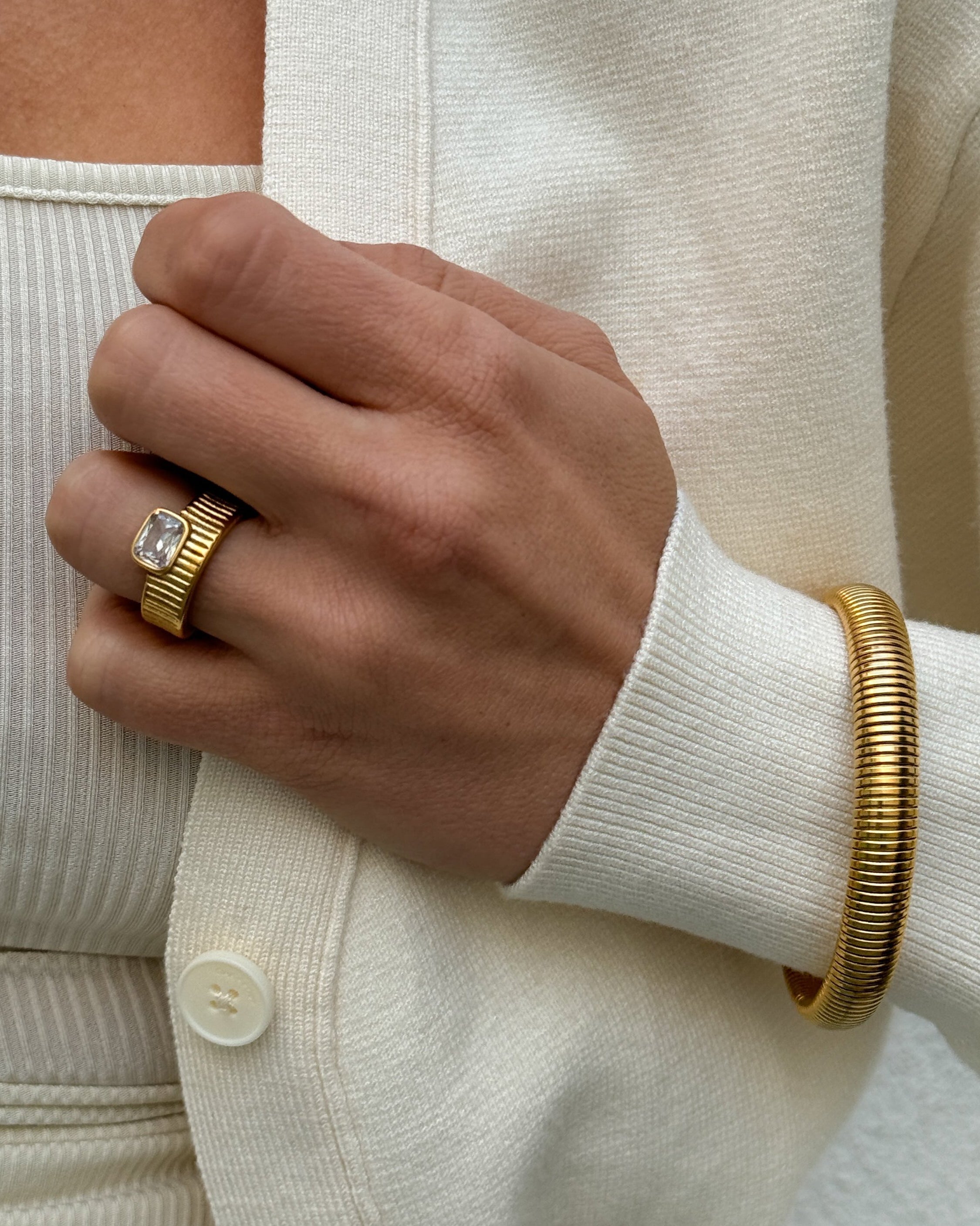 Hand wearing a gold ring and bracelet against a neutral background