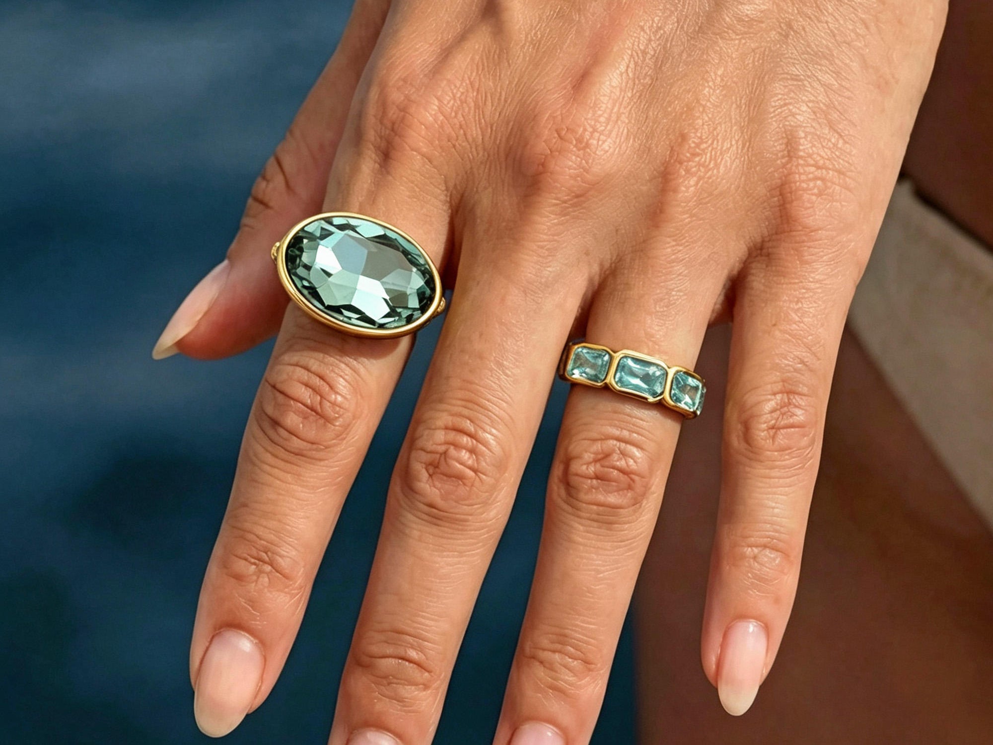Hand wearing two gold rings with green gemstones against a blurred natural background
