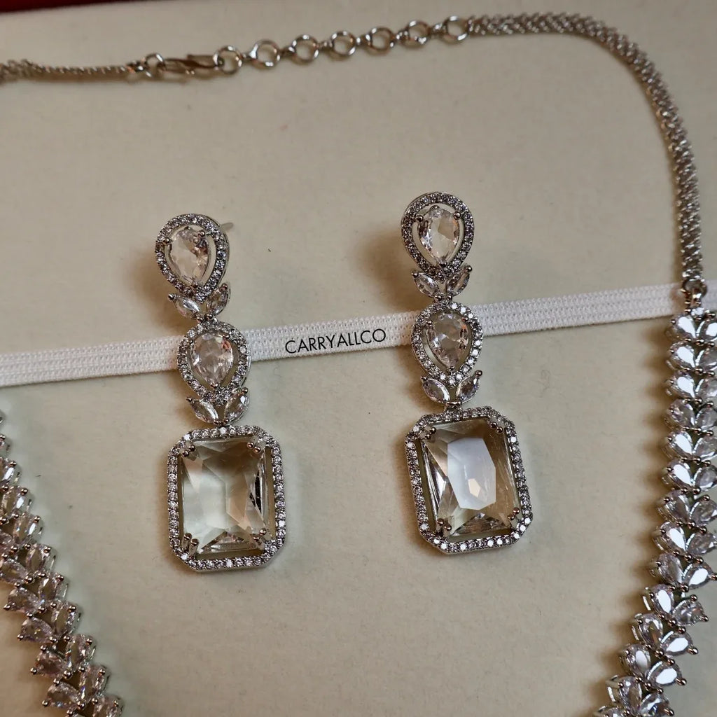 Pair of silver earrings with clear gemstones on a beige background with 'CARRYALLCO' branding.