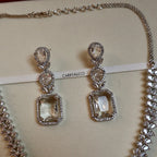 Pair of silver earrings with clear gemstones on a beige background with 'CARRYALLCO' branding.