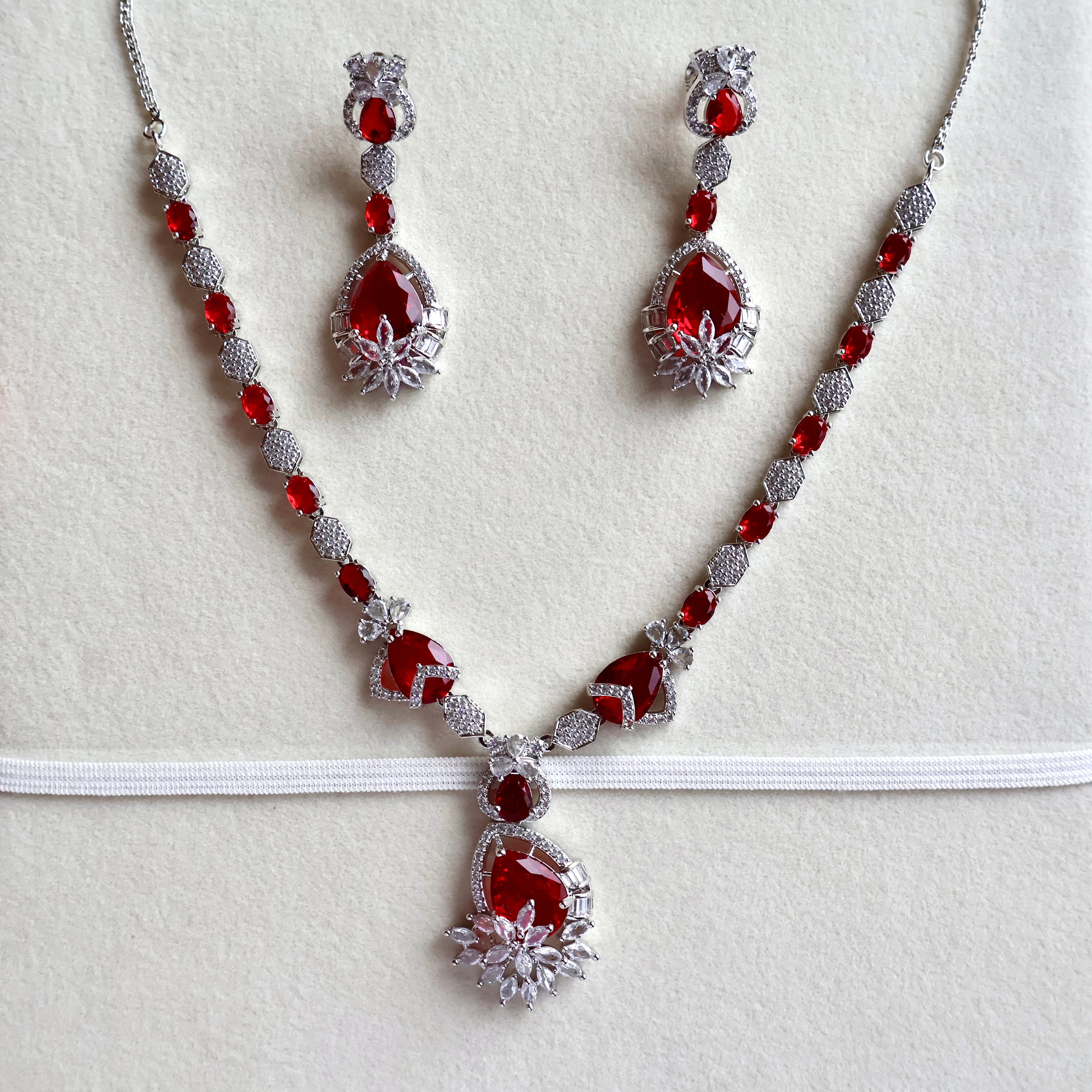 A red and silver jewelry set featuring a necklace, matching earrings, and a headpiece displayed on a textured white background.