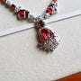 A close-up of a silver-toned necklace with prominent red gemstones and clear crystal accents, resting on a cream-colored textured surface with a white elastic band behind it.