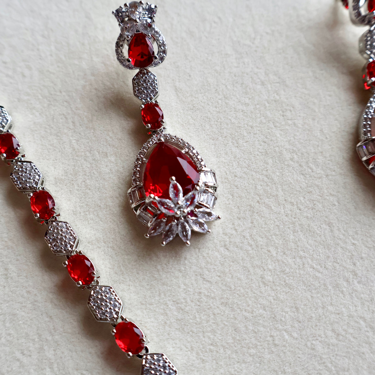 A close-up of a silver-toned jewelry set featuring a teardrop-shaped drop earring and a matching beaded bracelet, both adorned with vibrant red gemstones and clear crystals on a textured off-white background.
