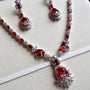 Silver-tone jewelry set with red gemstones, including a necklace, matching earrings, and a white fabric ribbon laid diagonally across a light beige background.