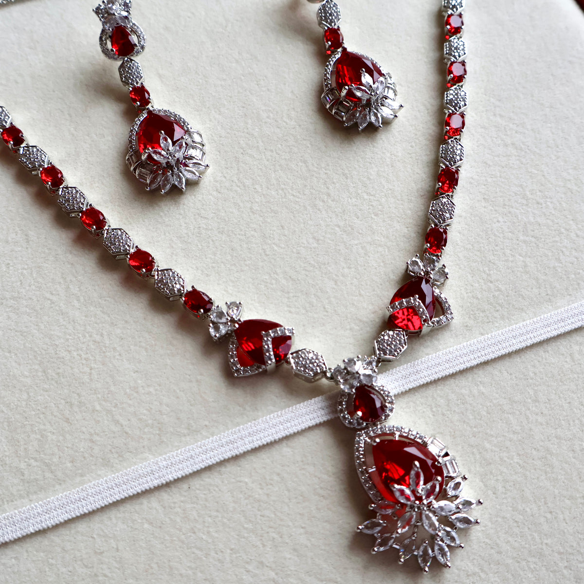 Silver-tone jewelry set with red gemstones, including a necklace, matching earrings, and a white fabric ribbon laid diagonally across a light beige background.