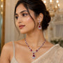A woman wearing a matching jewelry set featuring a silver-toned necklace and earring with deep red teardrop-shaped stones and clear crystal-like accents, styled with a beige embellished sari in an elegant indoor setting.
