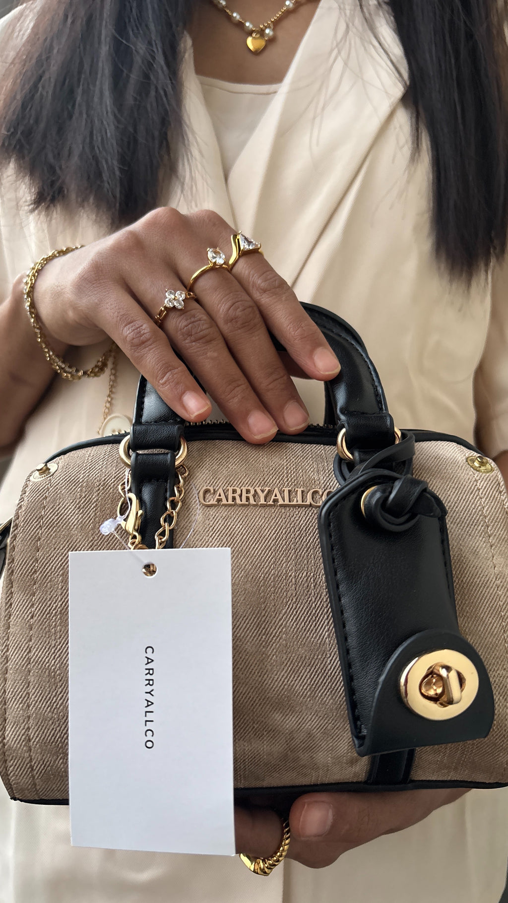 Person holding a beige handbag with a gold handle and 'CARRIAGELO' tag, wearing gold jewelry.