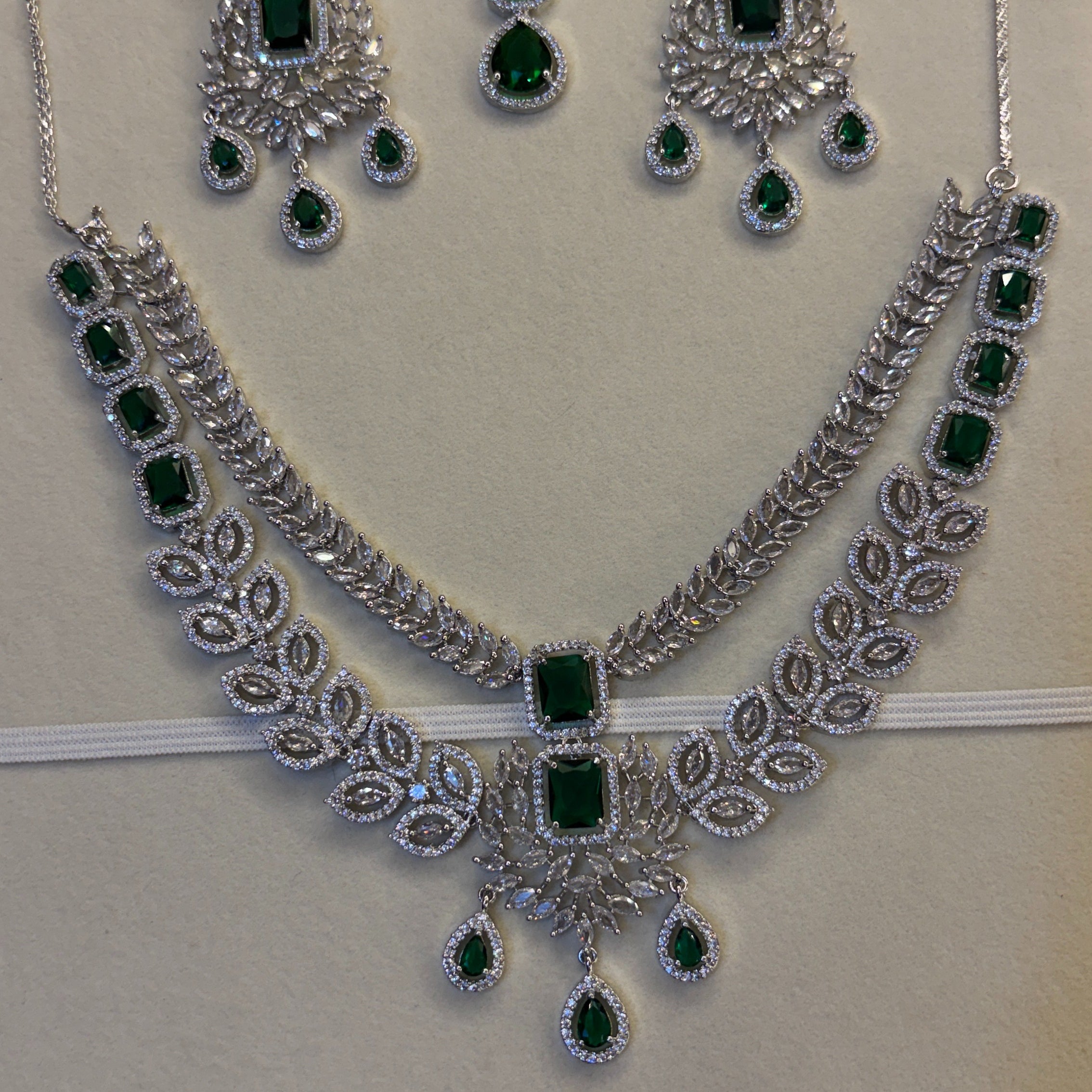 A silver-tone jewelry set displayed on a cream jewelry box insert, featuring a necklace, matching earrings, and a pair of dangling stones. The set is decorated with multiple rectangular and teardrop-shaped green emerald-cut stones, set against a dark blue box base with the brand name 'CRYALLCO' partially visible.
