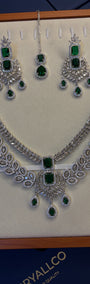 A silver-tone jewelry set displayed on a cream jewelry box insert, featuring a necklace, matching earrings, and a pair of dangling stones. The set is decorated with multiple rectangular and teardrop-shaped green emerald-cut stones, set against a dark blue box base with the brand name 'CRYALLCO' partially visible.