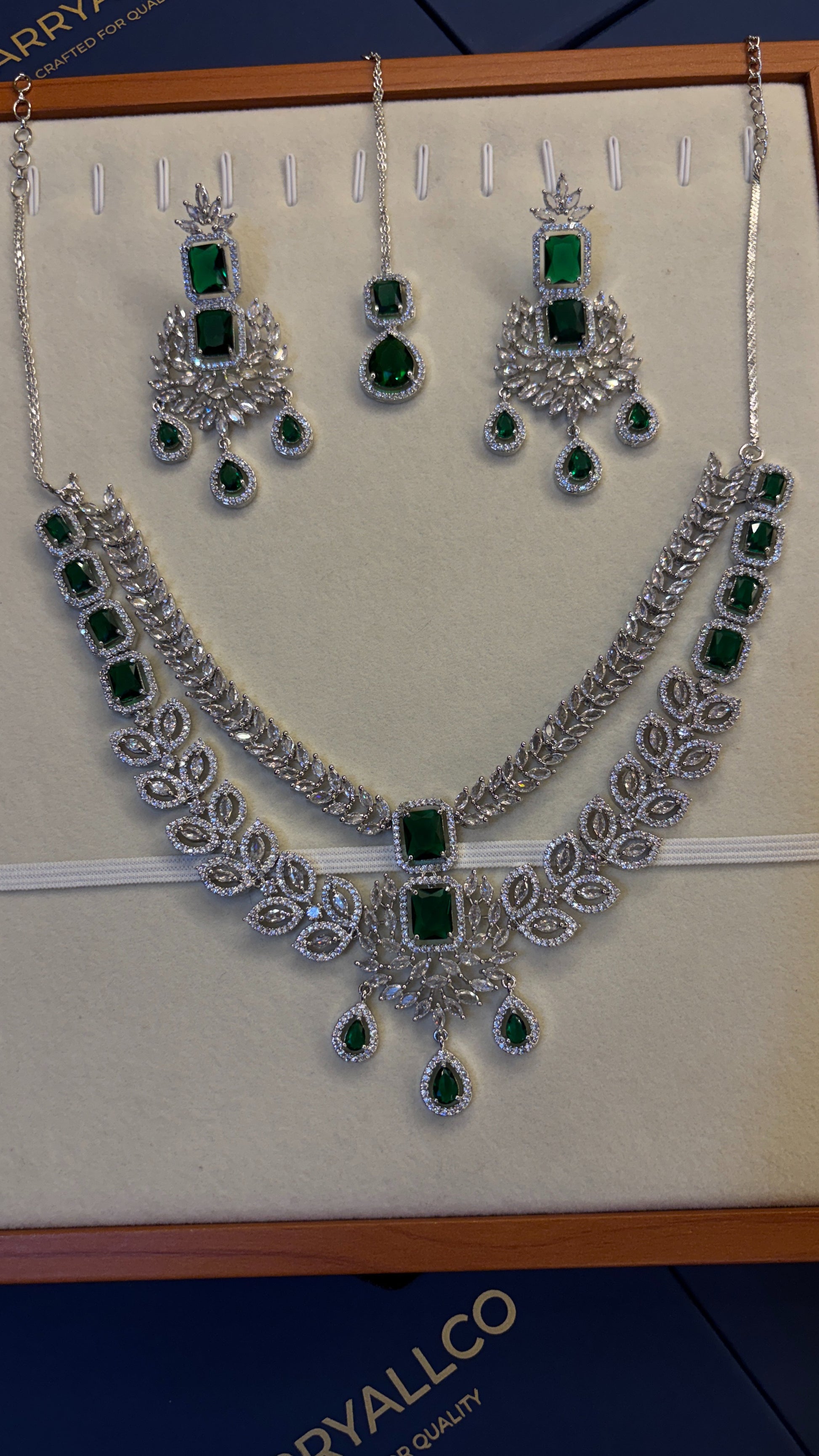 A silver-tone jewelry set displayed on a cream jewelry box insert, featuring a necklace, matching earrings, and a pair of dangling stones. The set is decorated with multiple rectangular and teardrop-shaped green emerald-cut stones, set against a dark blue box base with the brand name 'CRYALLCO' partially visible.