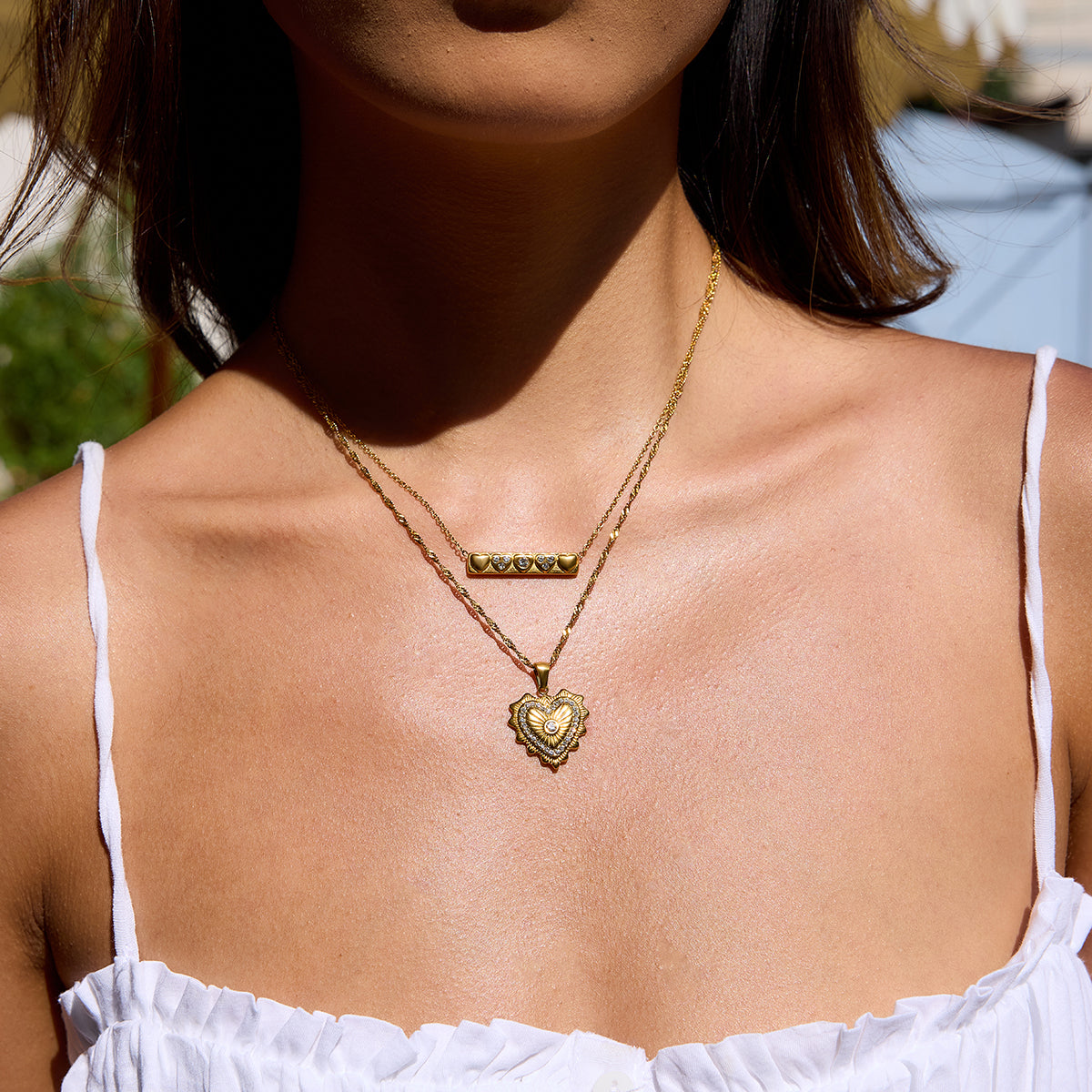 Close-up of a person wearing a gold necklace with a heart pendant, outdoors.