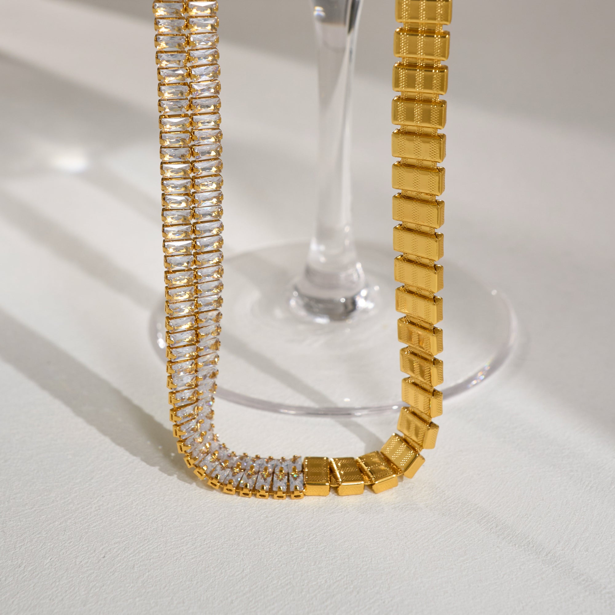 Gold and crystal bracelet on a white surface with a glass object in the background