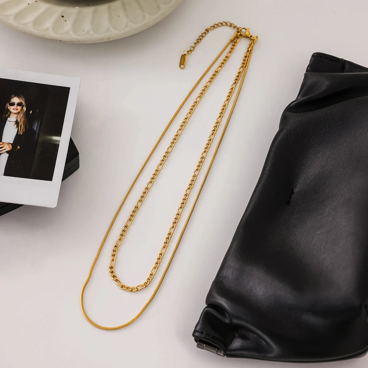 Gold chain necklace on a white surface with a black leather bag and photo album.
