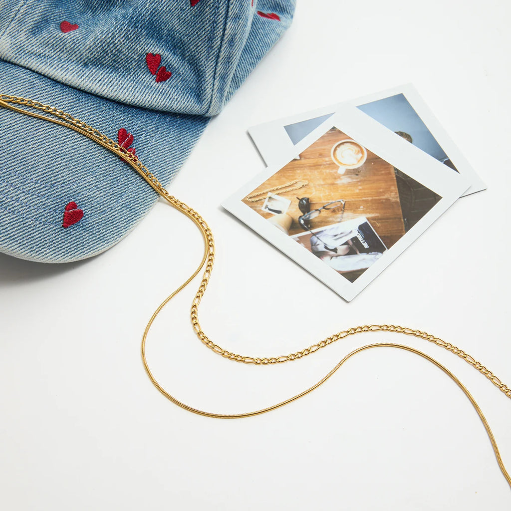 Gold chain necklace on a white surface with a denim cap and Polaroid photos.