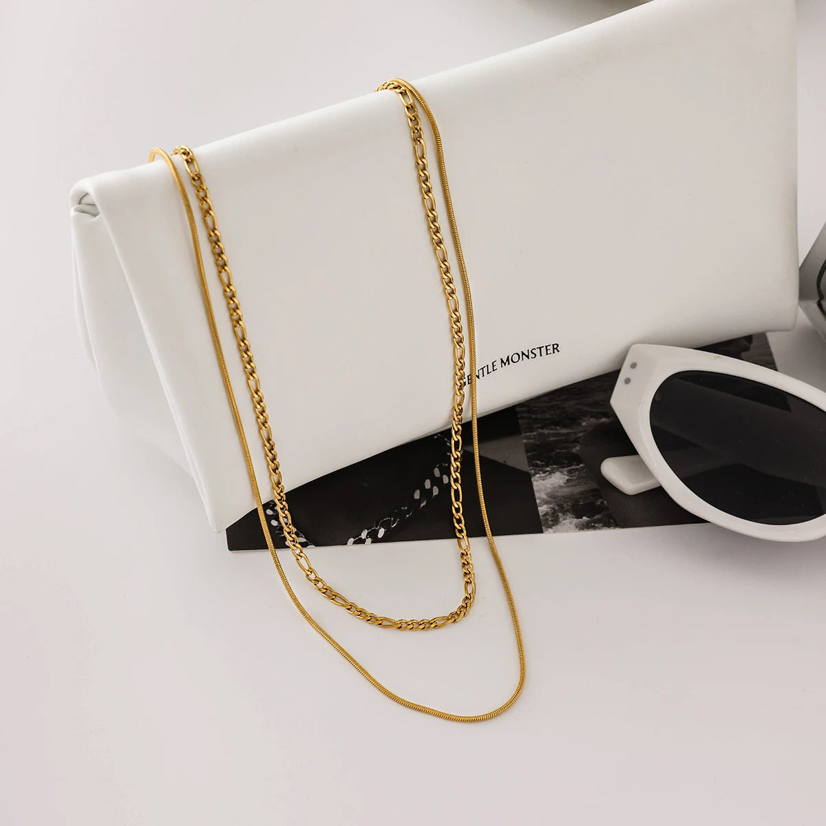 Gold chain necklace draped over a white clutch with sunglasses on a light gray background