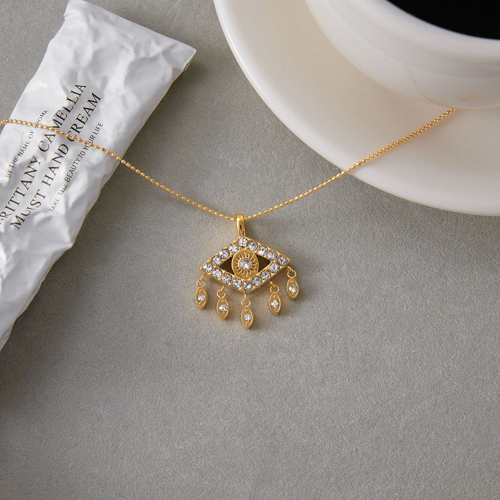 Gold necklace with an eye-shaped pendant on a gray surface next to a white cup.
