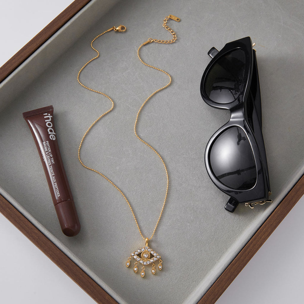 Gold necklace, tube of lipstick, and black sunglasses on a gray surface