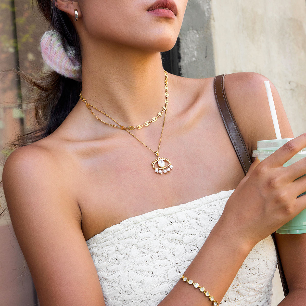 Woman wearing a gold necklace and bracelet, holding a drink.