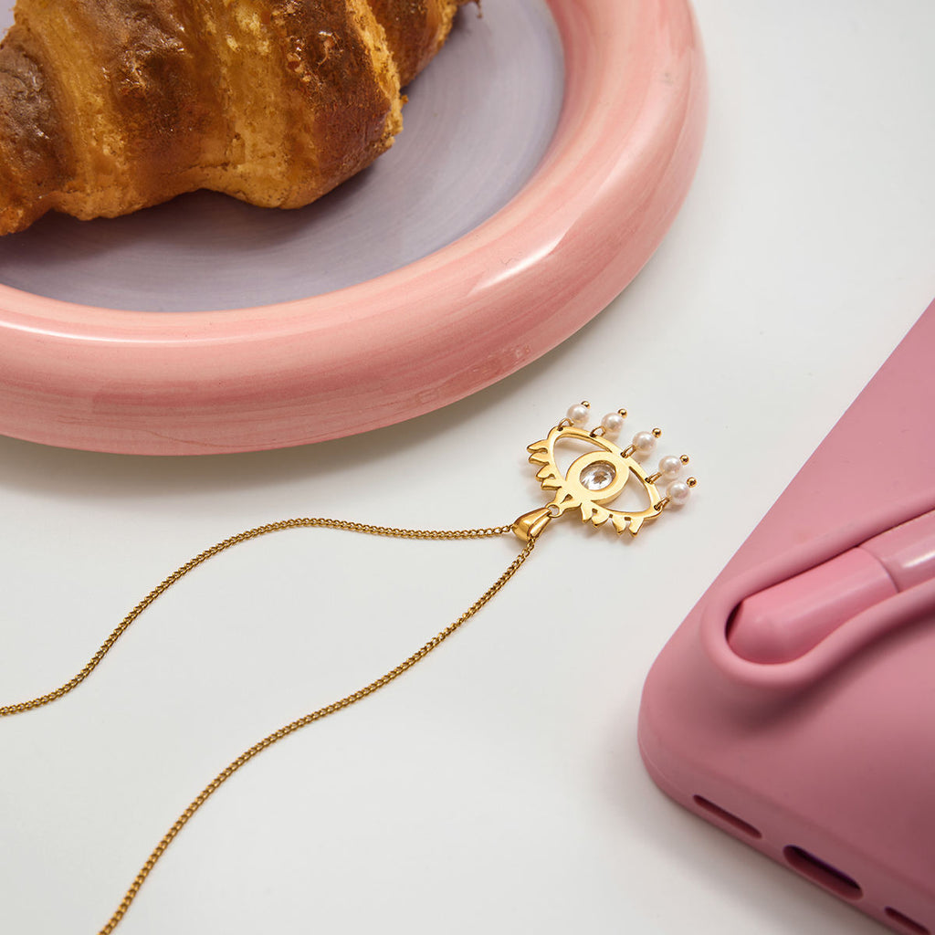 Gold phone charm with chain next to a pink phone case and croissant on a white surface