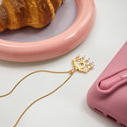 Gold phone charm with chain next to a pink phone case and croissant on a white surface
