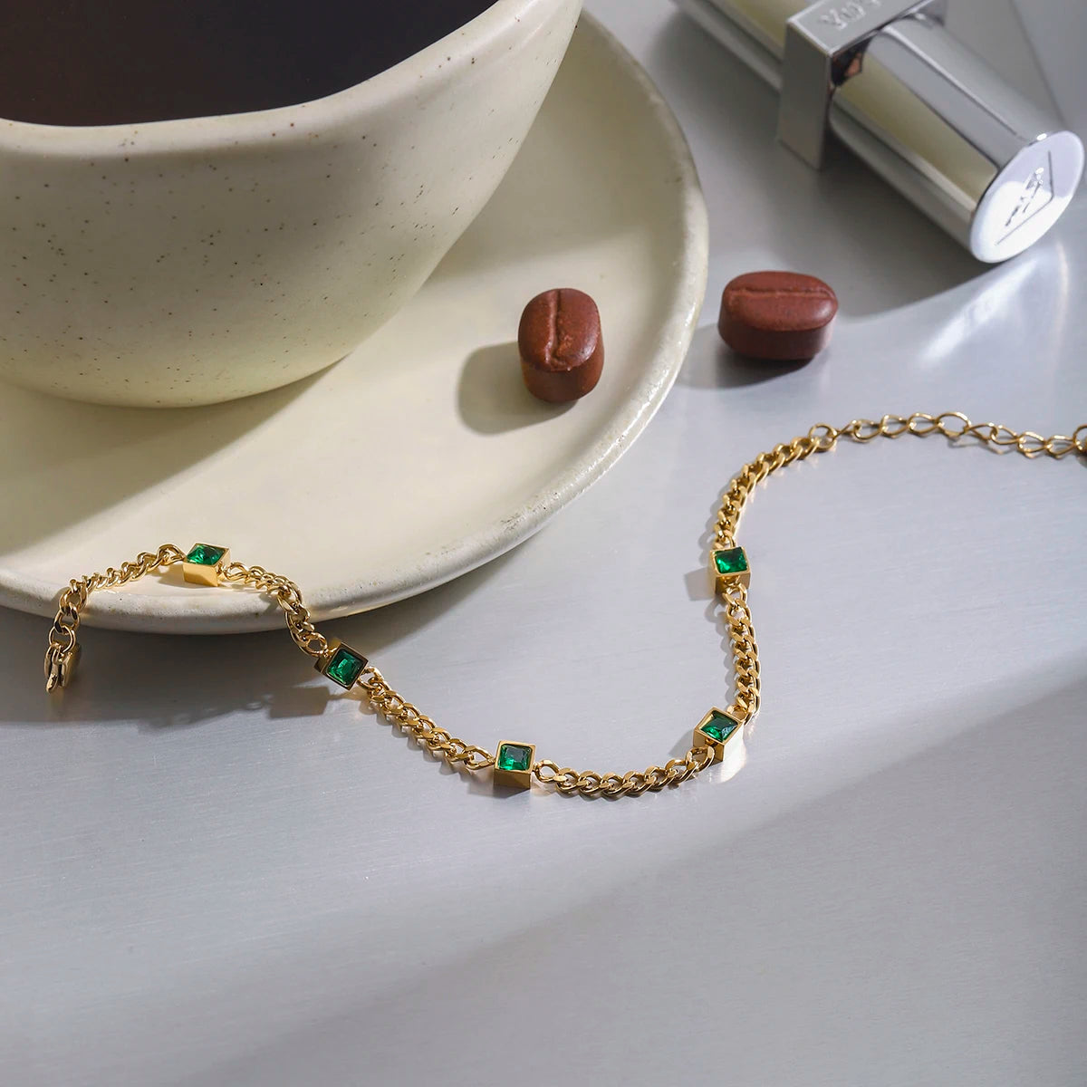 Gold bracelet with green gemstones on a white surface next to a cup and saucer.