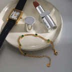 Gold watch, silver lipstick, gold bracelet with green stones on a white plate.