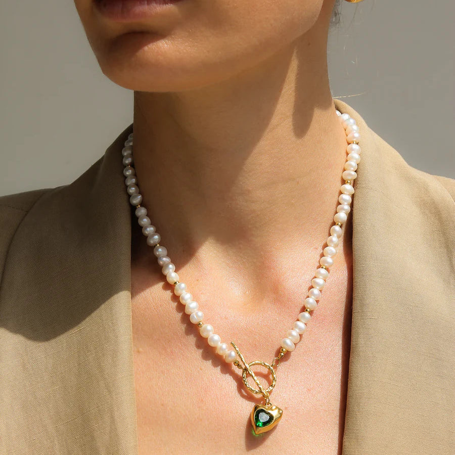Close-up of a person wearing a pearl necklace with a gold heart pendant and gold earrings.