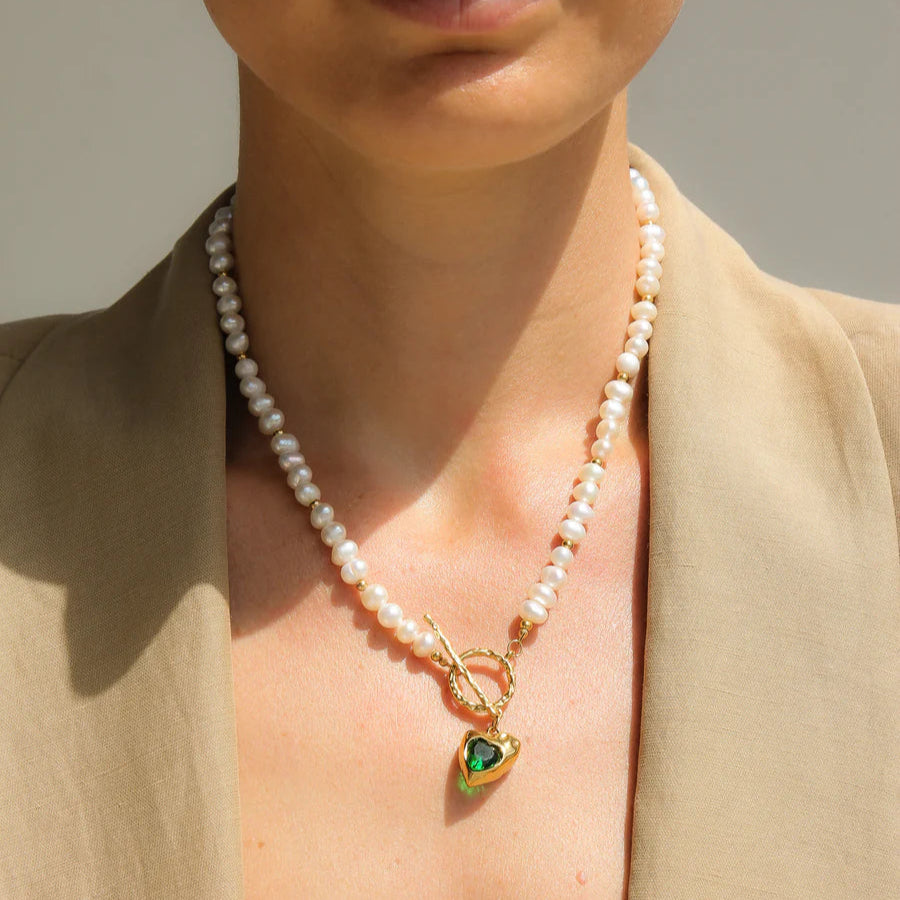 Woman wearing a pearl necklace with a green heart pendant against a neutral background