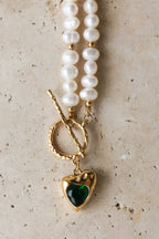 Pearl necklace with a gold clasp and green heart pendant on a textured beige surface