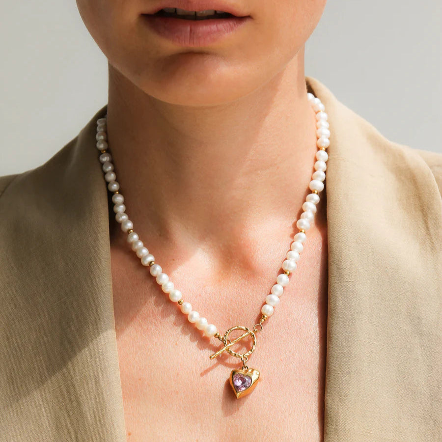 Pearl necklace with a heart charm worn by a person in a beige outfit.