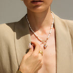 Woman wearing a pearl necklace with a heart pendant, gold earrings, and bracelet against a neutral background.