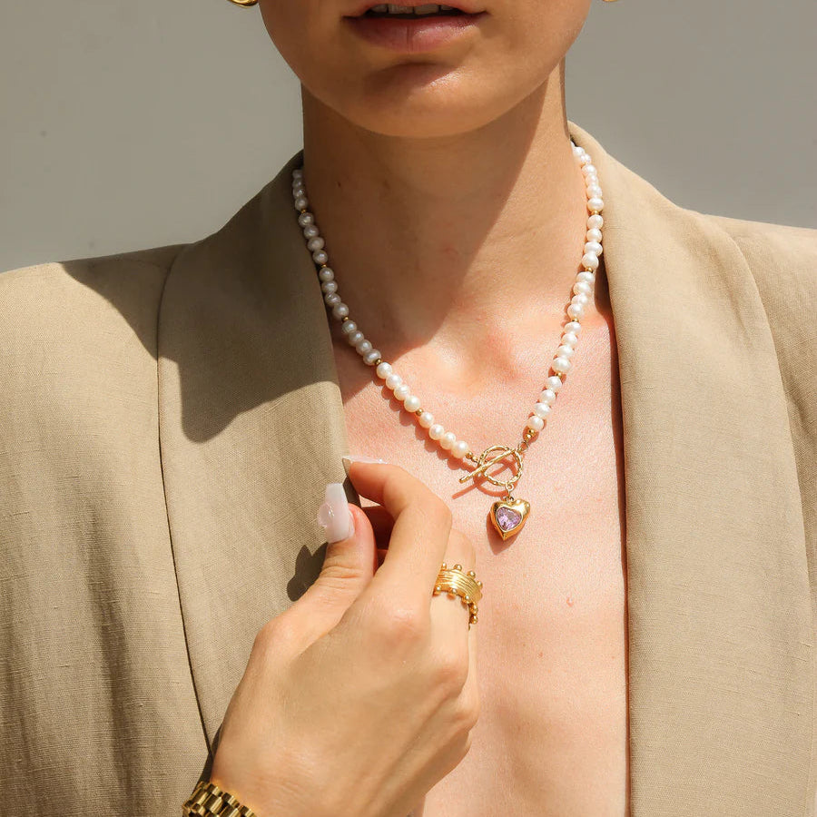 Woman wearing a pearl necklace with a heart pendant, gold earrings, and bracelet against a neutral background.
