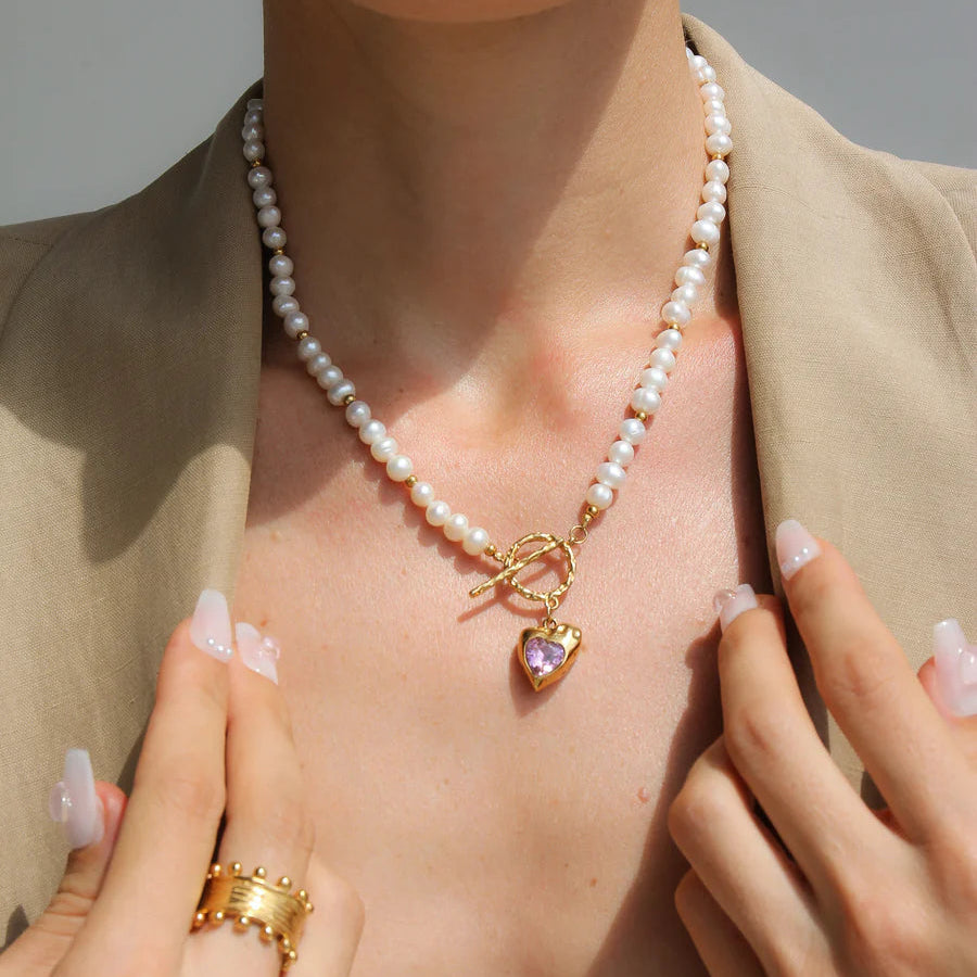 Person wearing a pearl necklace with a heart-shaped pendant, gold ring, and earrings against a neutral background.