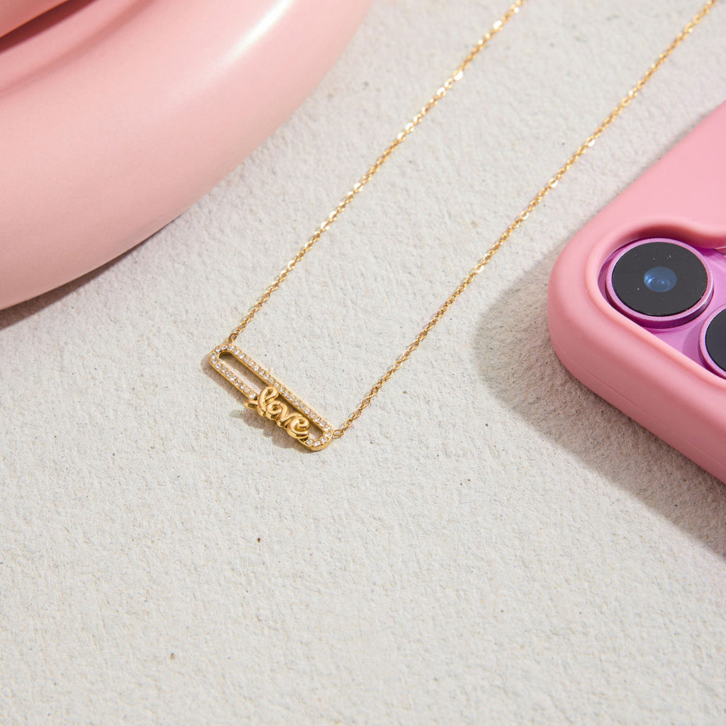 Gold necklace with 'love' pendant on a textured surface with pink phone case and pink object in the background
