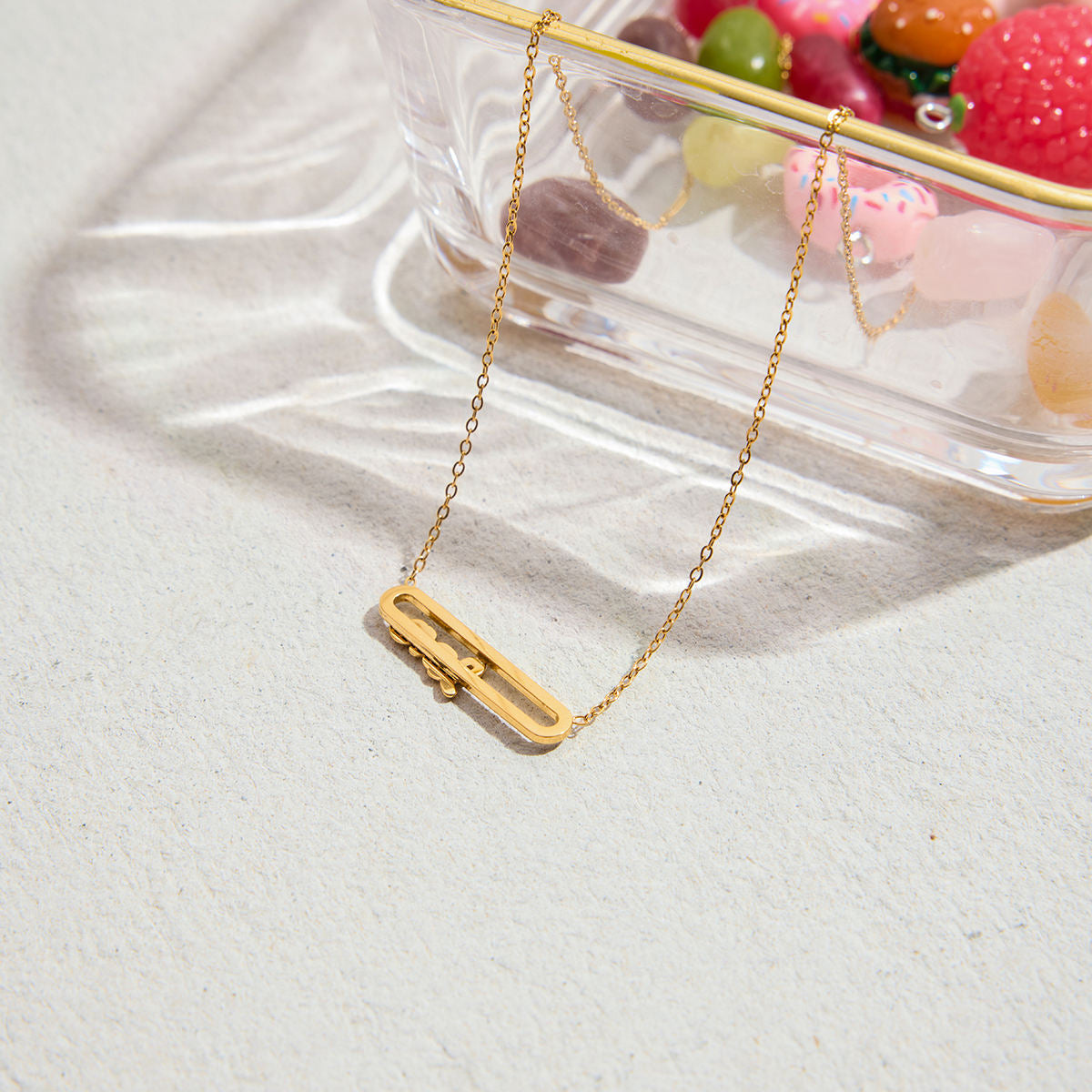 Gold necklace with a rectangular pendant on a light surface with colorful objects in the background