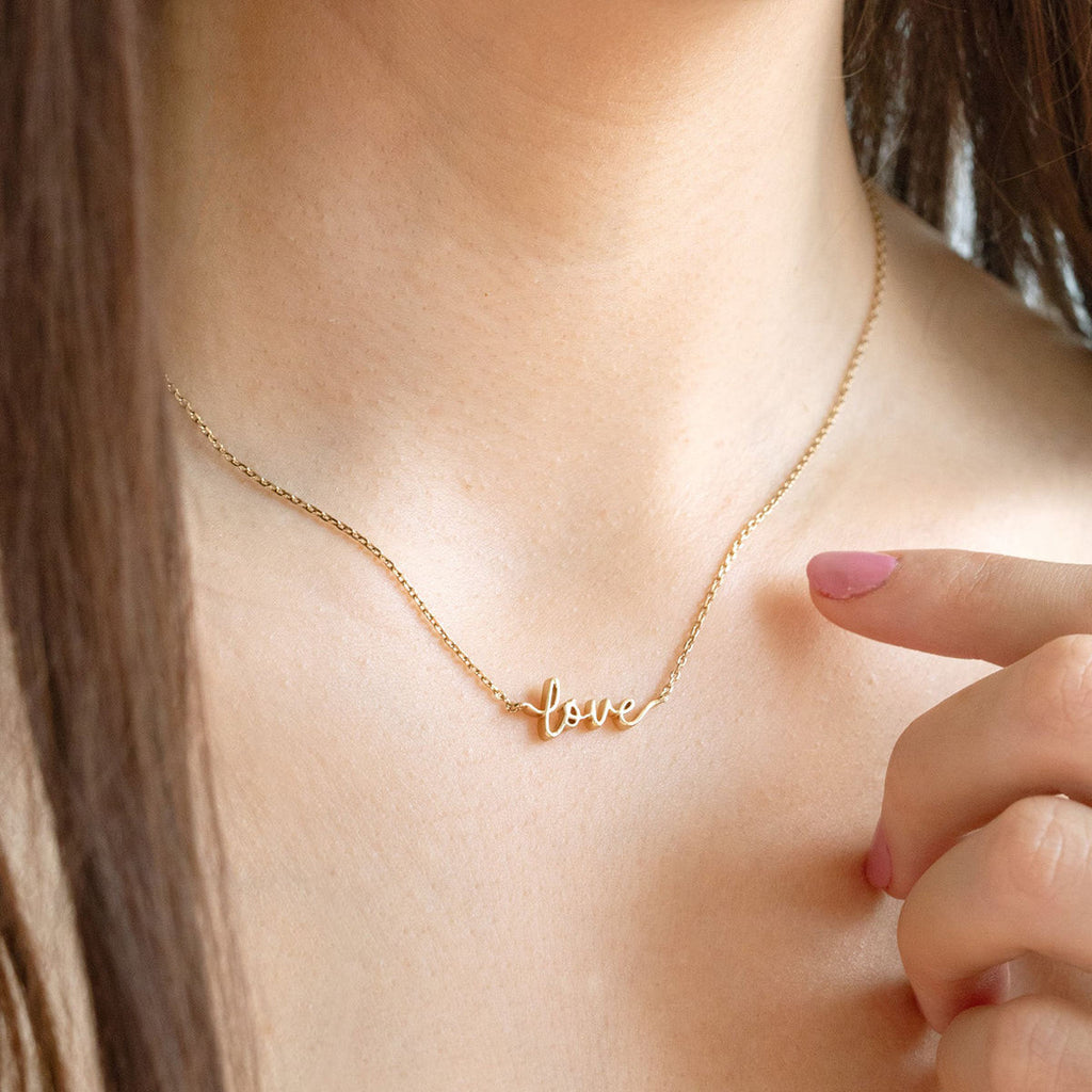 Gold 'love' necklace on a person's neck with a hand pointing to it.