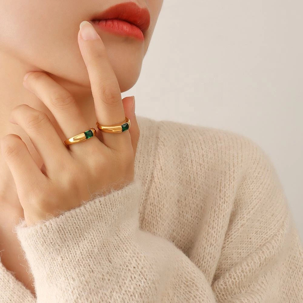 Close-up of a hand wearing two gold rings with green gemstones, against a neutral background.