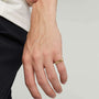 A close-up of a person's hand wearing a minimalist gold band ring, with a white shirt and dark trousers visible against a light gray background.