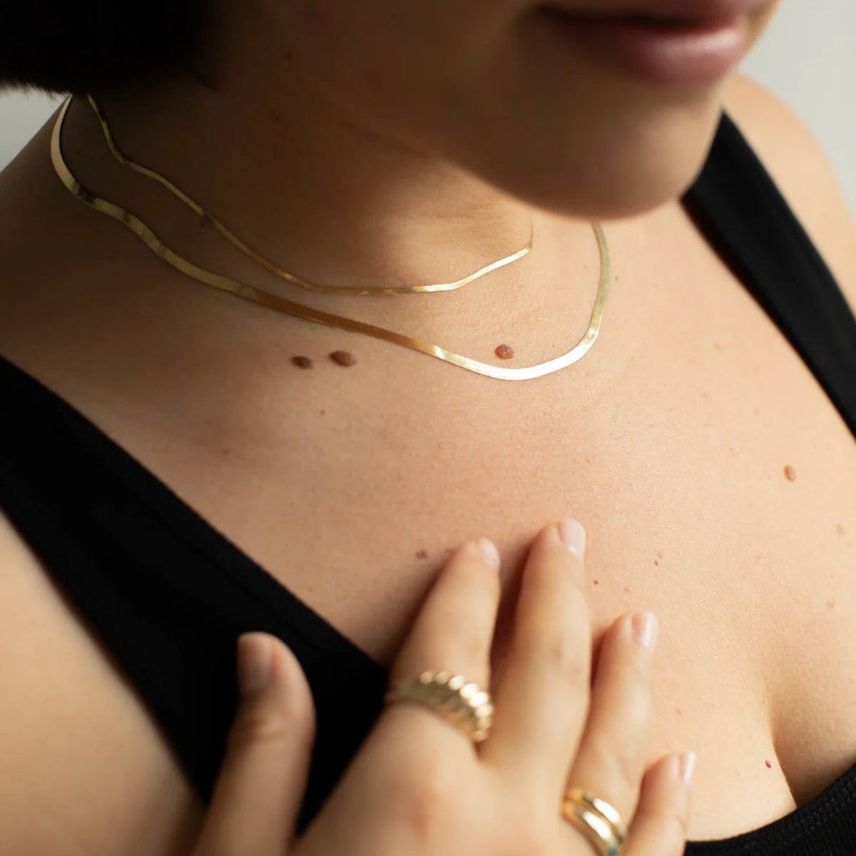 A close-up lifestyle shot of a model wearing two delicate gold chain necklaces layered on her neck, along with gold rings, against a black top.