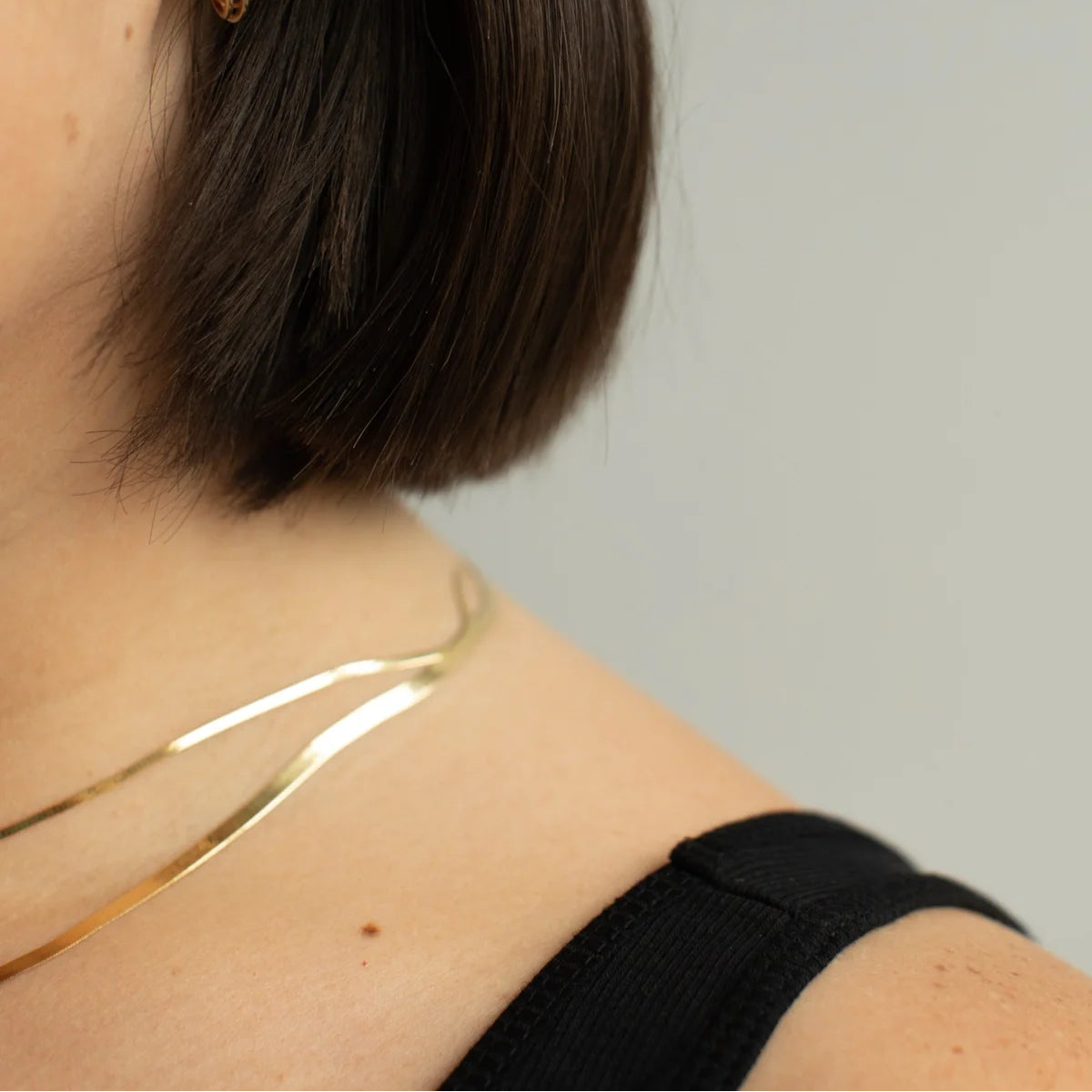 Close-up of a person's ear wearing a small gold hoop earring, with dark hair, a black top strap, and a thin gold necklace against a light gray background.