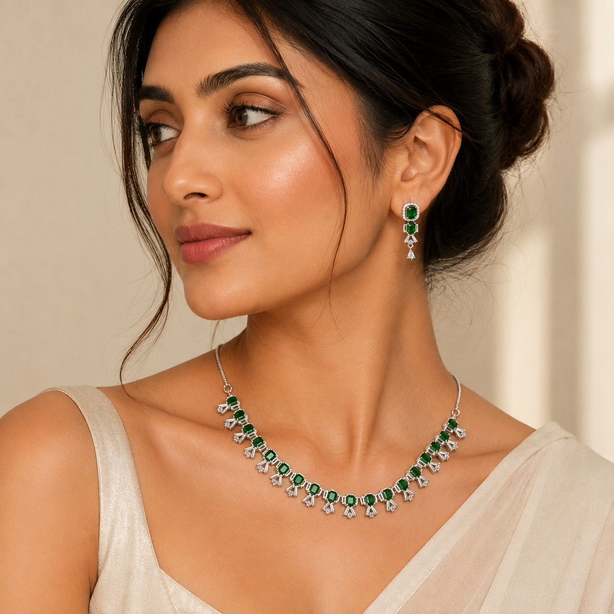 A woman wearing a silver-toned necklace and matching drop earring, both decorated with round green stones and clear teardrop-shaped crystals, shown against a soft beige background.
