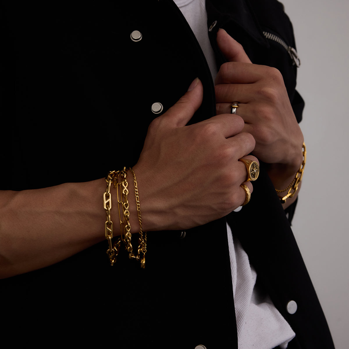 Close-up of hands with gold jewelry on a black jacket against a neutral background