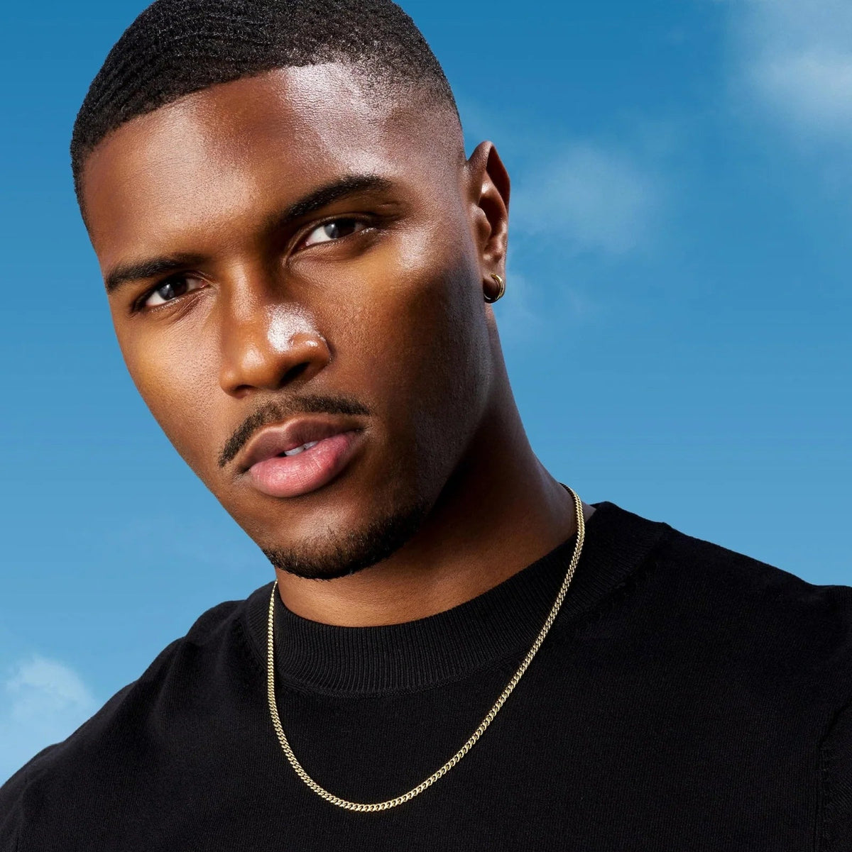 A close-up portrait of a man wearing a delicate gold chain necklace over a black crewneck shirt, set against a bright blue sky background.